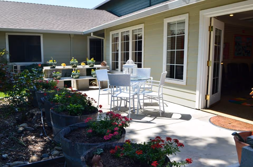 Outdoor patio area at Sundial Assisted Living with a white metal table and four chairs on a concrete surface. Several large planters with red and green flowering plants line the edge of the patio. The building exterior is light yellow with white trim around windows and a door leading inside. Additional flower pots with yellow flowers are arranged on a raised platform near the building.