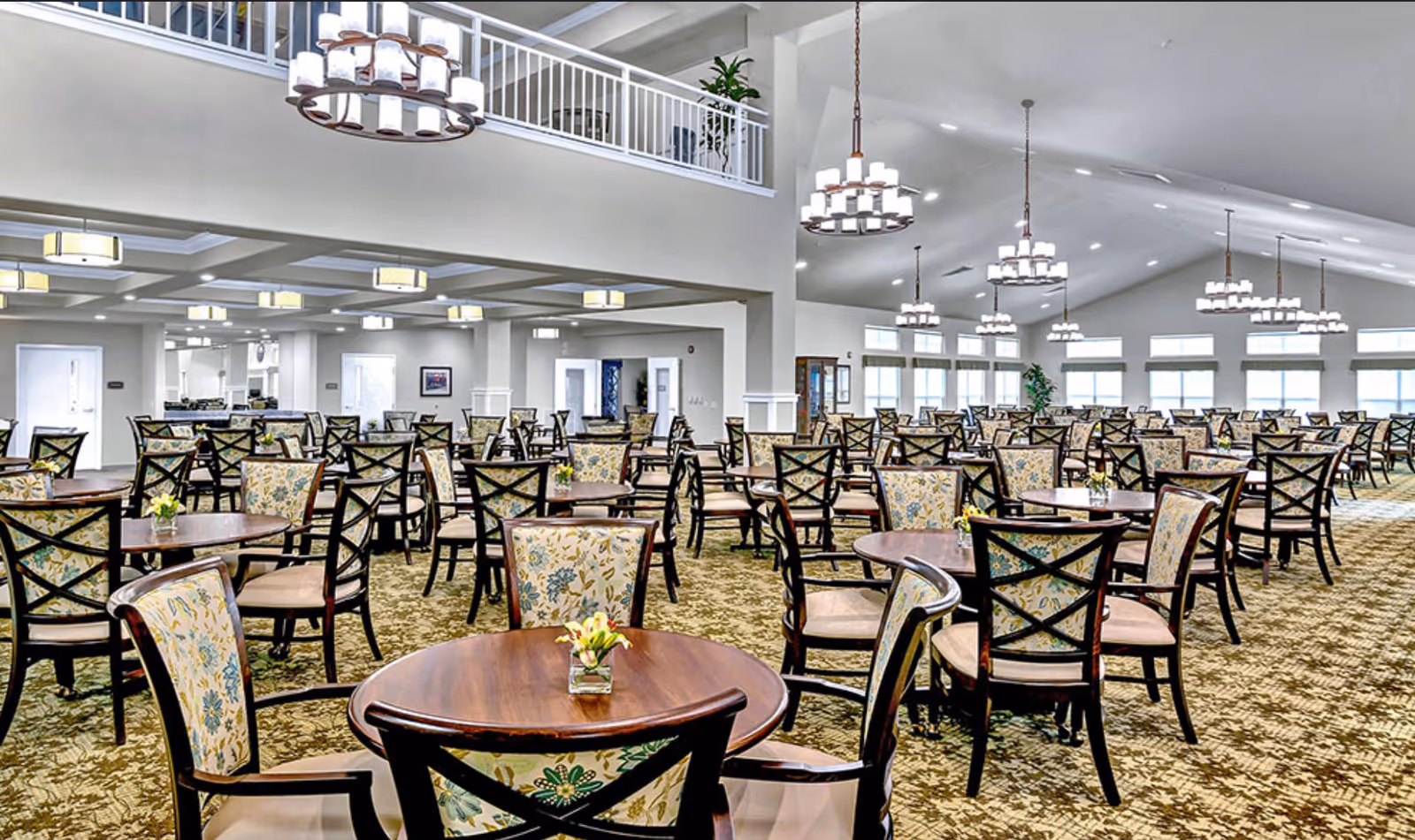 Spacious dining room with multiple round wooden tables and chairs featuring floral upholstery. The room has high ceilings with modern chandeliers and large windows allowing natural light to fill the space. The carpet has a patterned design, and small flower arrangements are placed on each table.