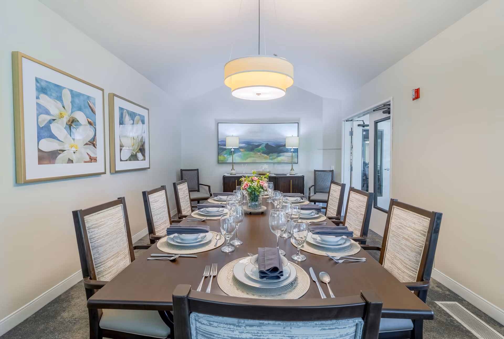 Formal dining room with a long table set for multiple guests, upholstered chairs, a pendant light, floral centerpiece, and framed artwork on the walls.