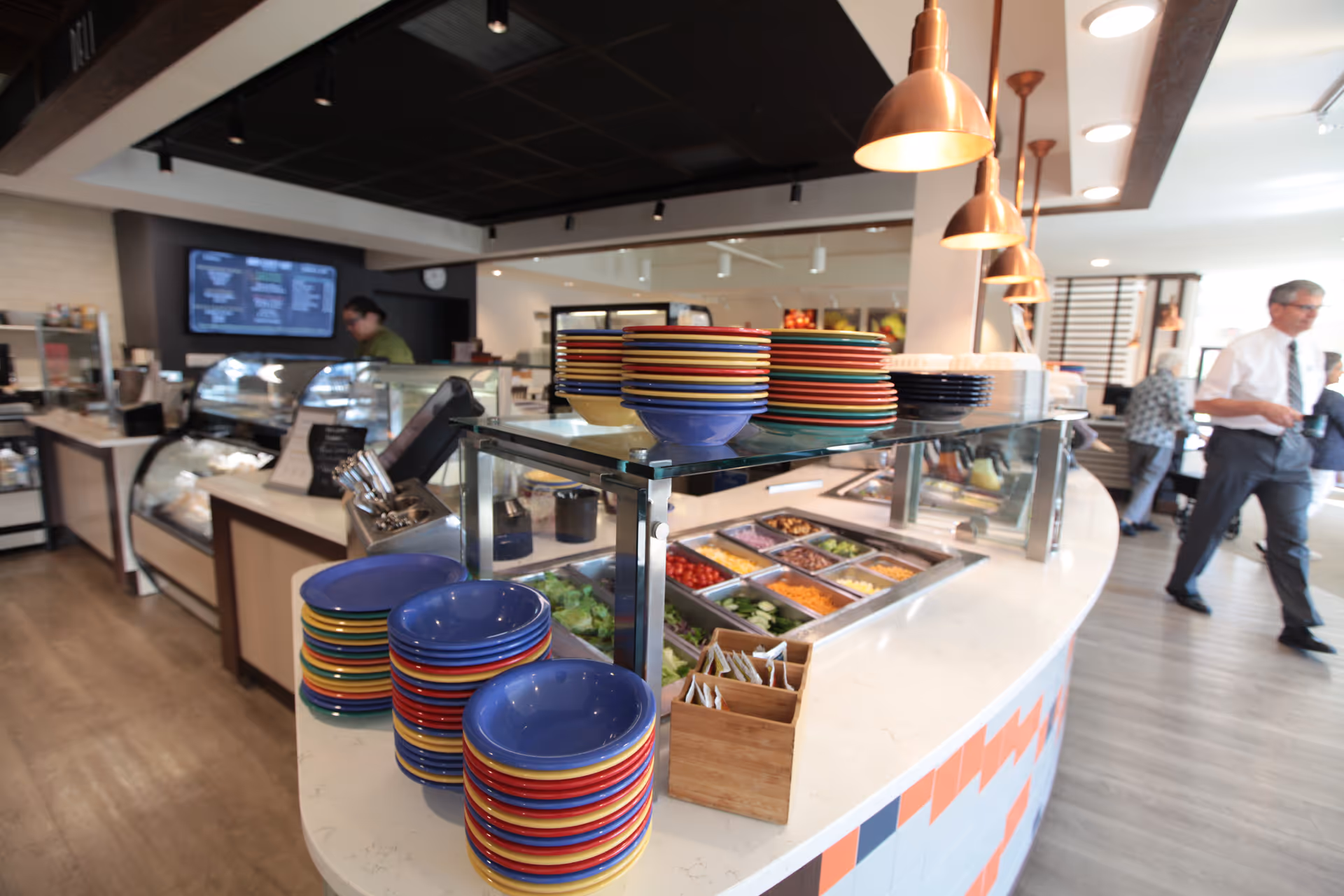 Interior view of a dining area with a salad bar counter featuring colorful stacked plates and bowls. Behind the counter, a person is working near refrigerated display cases. Several pendant lights hang from the ceiling, and people are seen walking in the background.