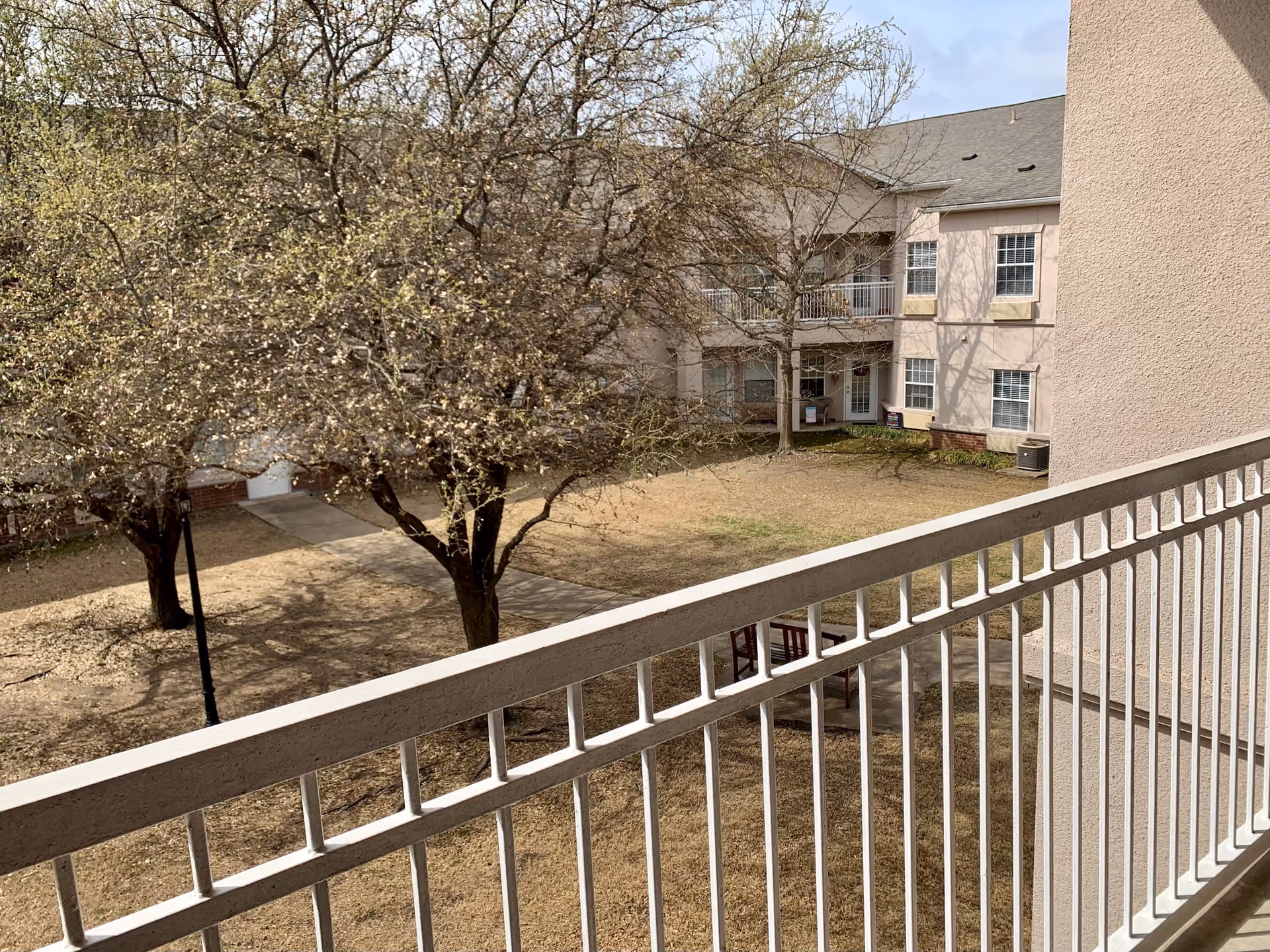 View from a balcony overlooking a courtyard with trees, a walkway, and the adjacent apartment building.