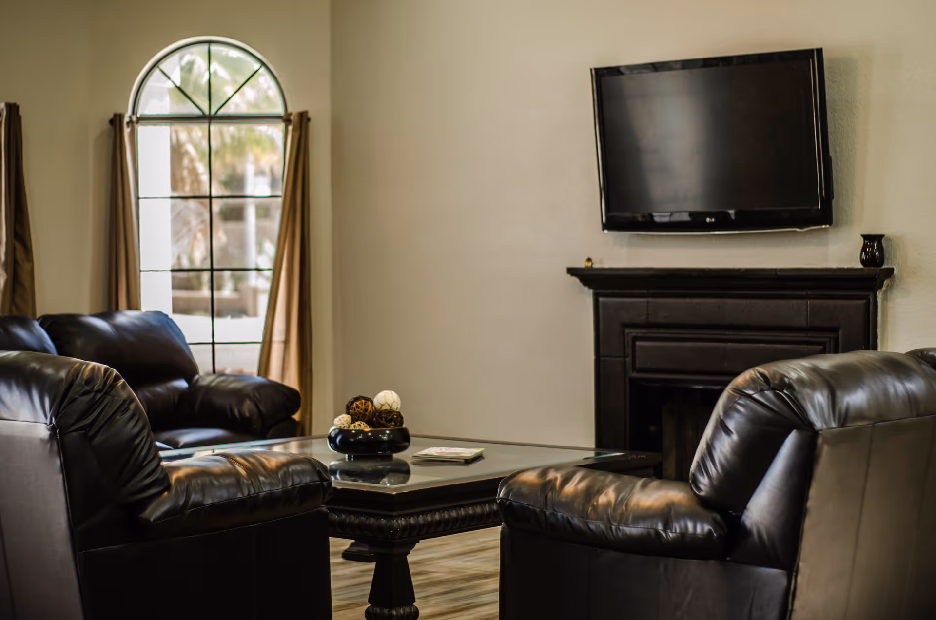 A cozy living room with two black leather armchairs and a matching sofa arranged around a glass-top coffee table. The table has a decorative bowl with assorted decorative balls and a small stack of magazines. A flat-screen TV is mounted above a dark fireplace on the wall. A large arched window with beige curtains allows natural light into the room.