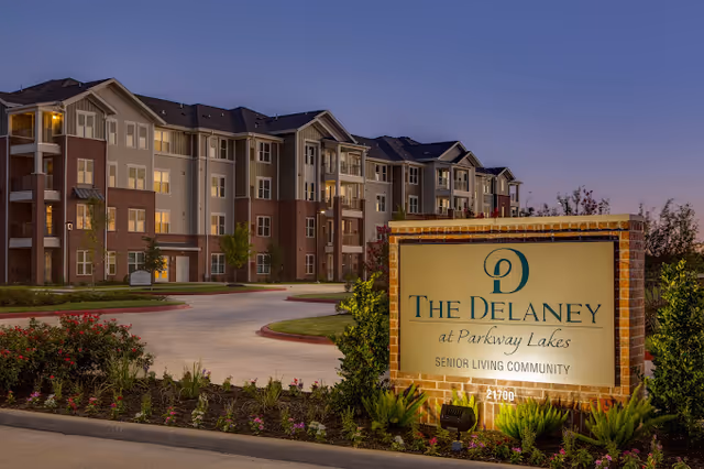 Exterior view of The Delaney at Parkway Lakes senior living community building during dusk, with a large illuminated sign in the foreground surrounded by landscaping and a driveway leading to the building entrance.