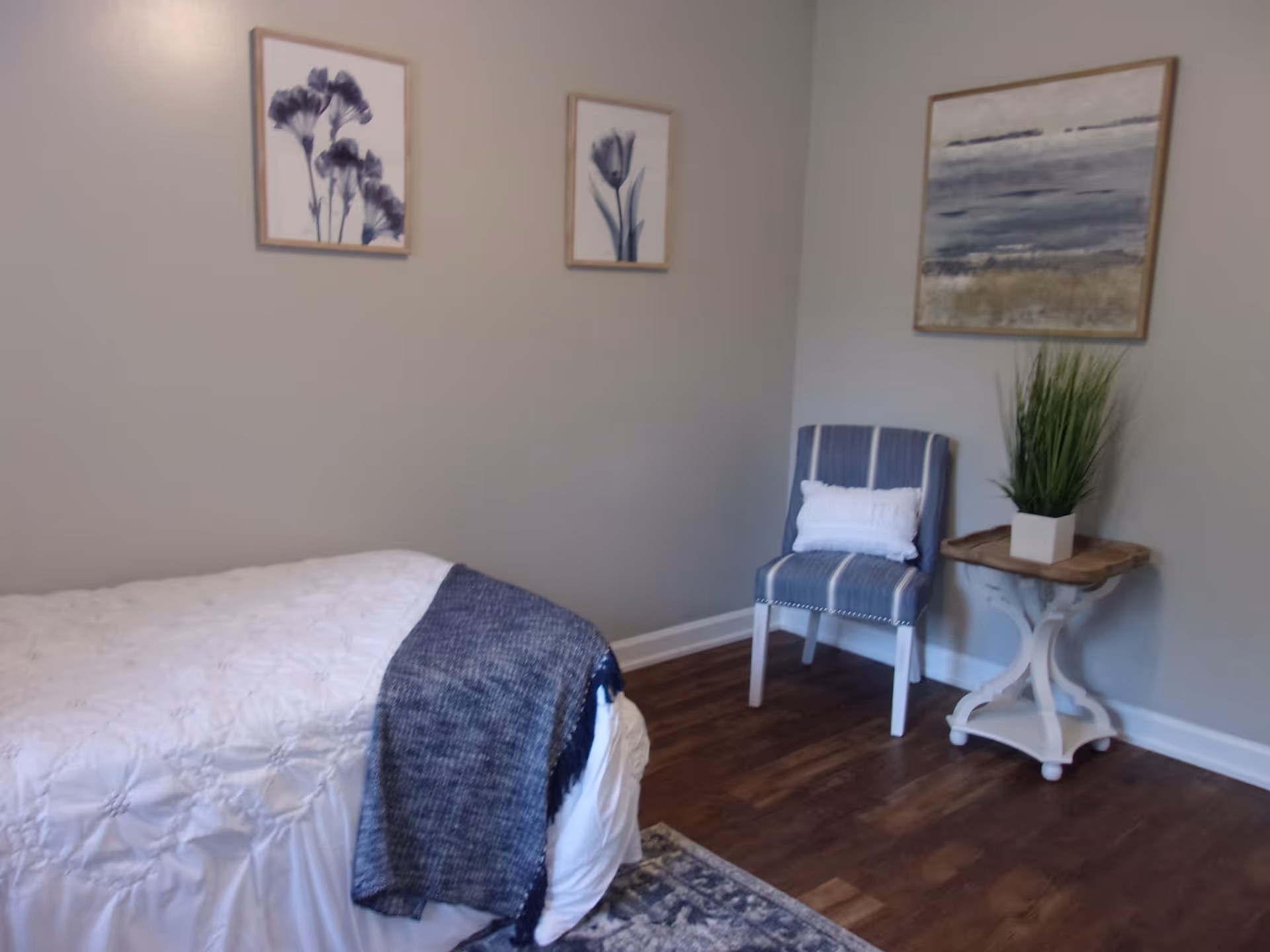 A bedroom with a bed covered in a white quilt and a dark blue throw blanket. There is a blue and white striped chair with a white pillow next to a small wooden side table holding a potted green plant. Three framed artworks hang on the light gray walls above the bed and chair. The floor is wooden with a patterned rug partially visible.