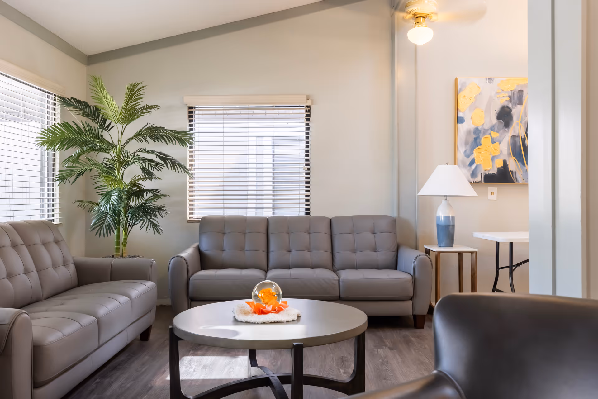 A cozy living room with two gray leather sofas and a matching armchair arranged around a round coffee table with a decorative centerpiece. There is a large green potted plant near the window with blinds, a side table with a blue and white lamp, and an abstract painting on the wall. The room has wood flooring and a ceiling fan with a light.
