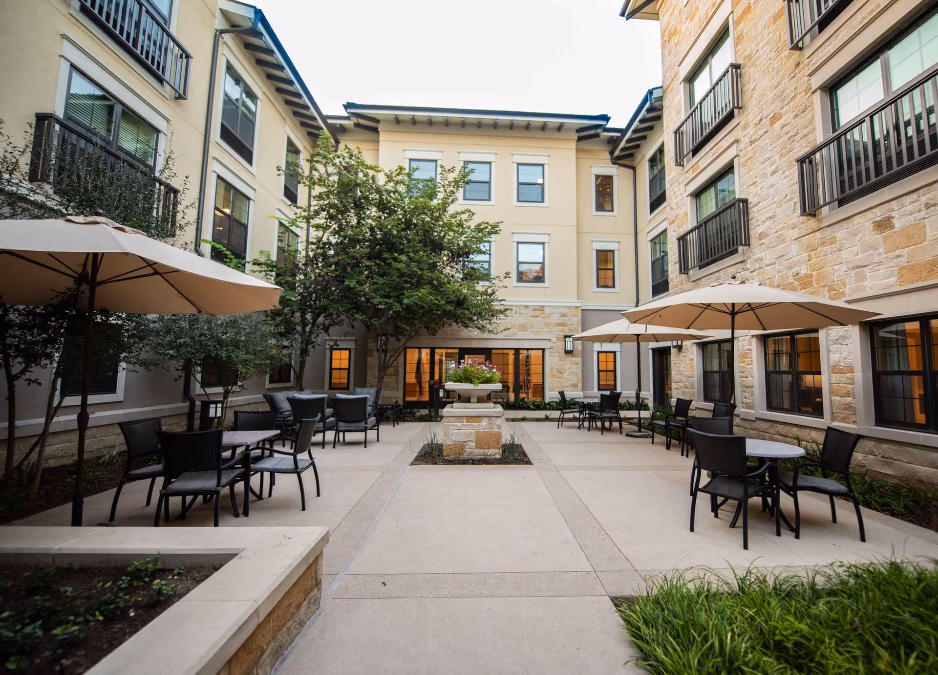 Outdoor courtyard area at Belmont Village Senior Living West Lake Hills featuring multiple tables with black chairs and large beige umbrellas. The courtyard is surrounded by a three-story building with beige and stone exterior walls, windows, and some greenery including trees and plants.