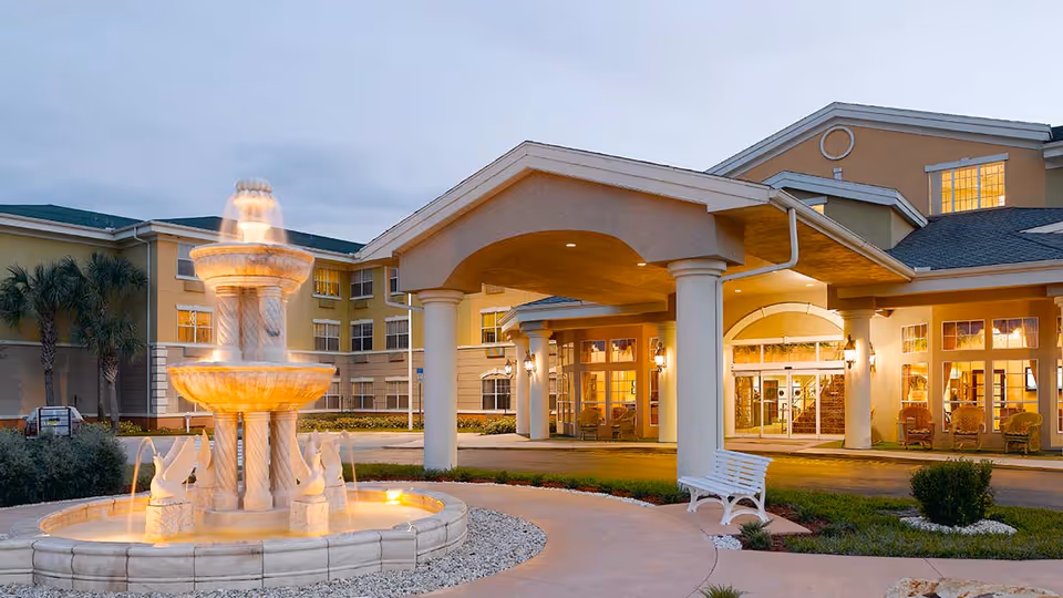 Entrance of Vitality Living Lake Forest facility at dusk with a large illuminated tiered fountain in the foreground, a white bench nearby, and the building's covered entryway with columns and large windows in the background.