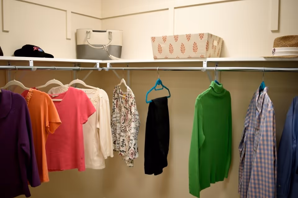 Interior view of a closet with various clothes hanging on a metal rod, including sweaters, shirts, and pants. On the shelf above, there are a beige and gray handbag, a patterned storage box, a hat, and a black cap.