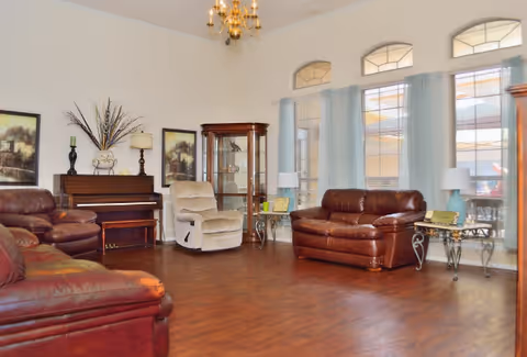 A spacious living room with wooden flooring featuring brown leather sofas, a beige recliner chair, a wooden piano with decorative items on top, a glass display cabinet, two side tables with lamps, and large windows with light blue curtains allowing natural light to enter.