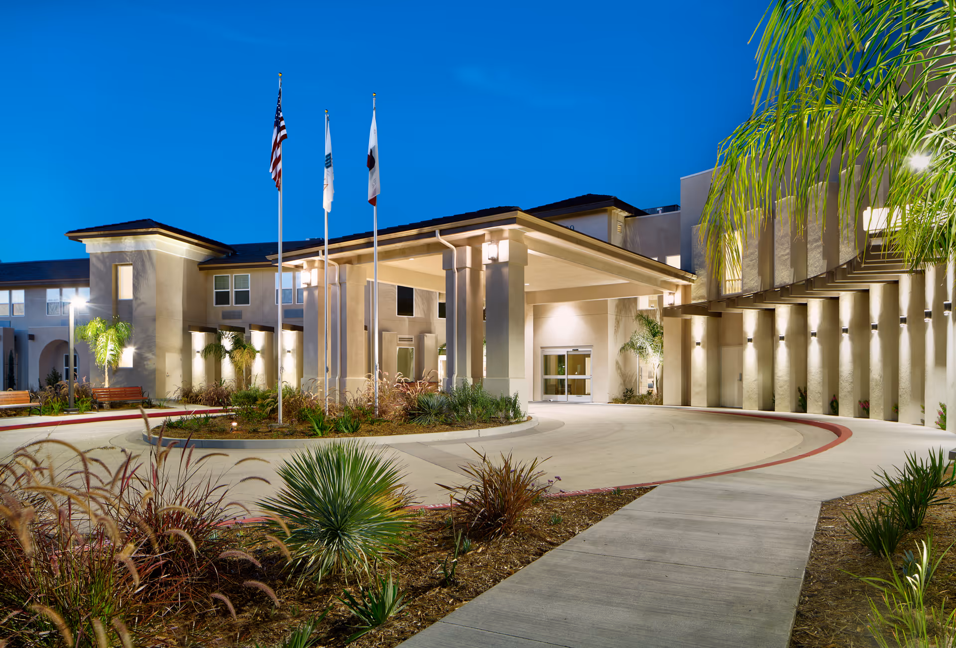 Exterior view of Ivy Park at Otay Ranch senior living facility at dusk, showing the entrance with a covered drop-off area, three flagpoles with flags, landscaped plants, and well-lit building facade.