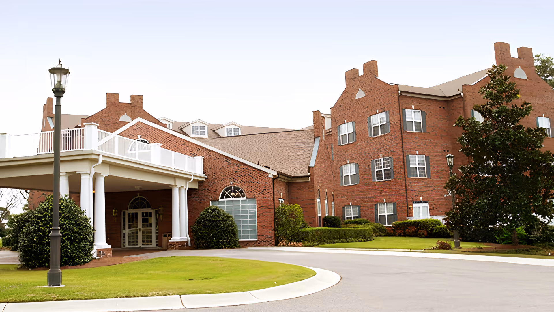 Red-brick building with a covered porte-cochère entrance, manicured lawn, and a lamppost in front.