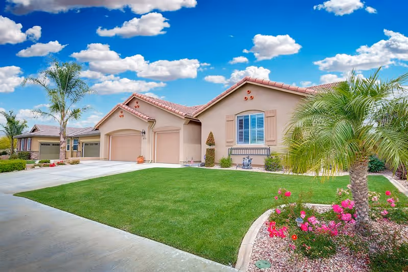 Exterior view of a single-story residential building with a well-maintained green lawn, palm trees, and colorful flowers under a blue sky with scattered clouds.