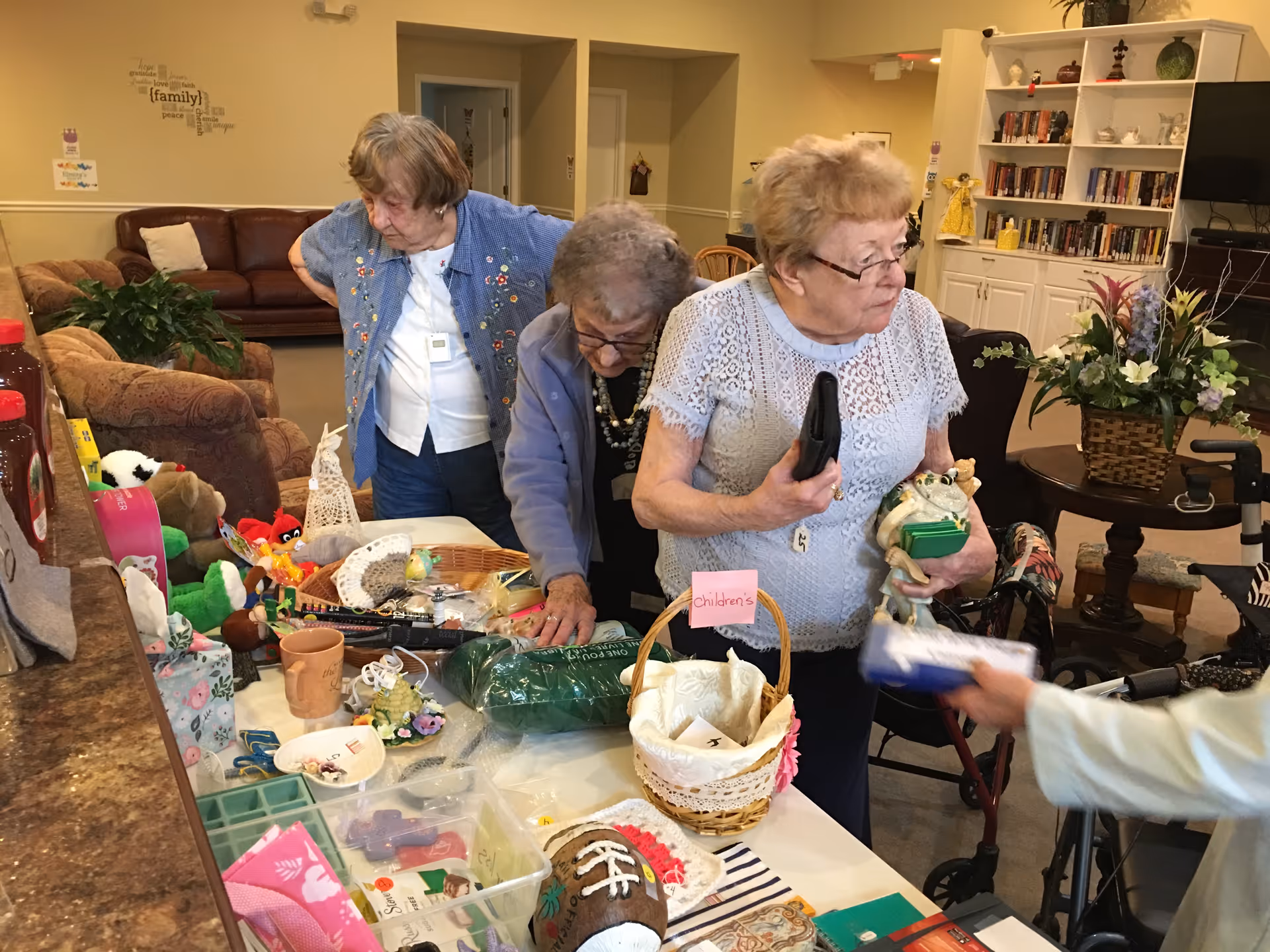 Three elderly women standing around a table filled with various small items and crafts in a cozy living room area. One woman is holding a figurine and a remote control, while another is looking closely at the items on the table. The room has comfortable seating, a bookshelf with books, and a floral arrangement on a side table.