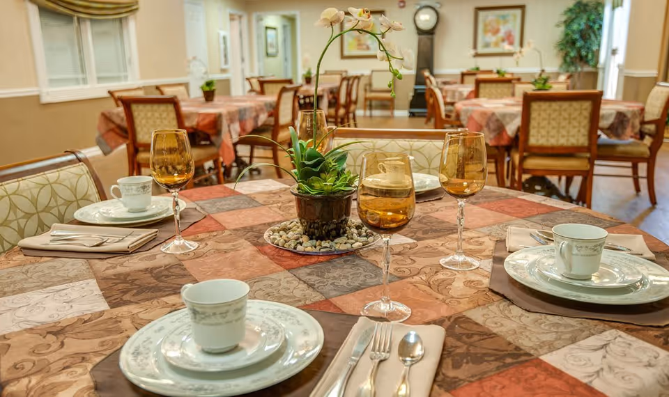 A dining room in a senior living facility with tables covered in patterned tablecloths. Each table is set with plates, cups, silverware, and amber-colored wine glasses. A potted orchid plant serves as a centerpiece on the table in the foreground. The room has warm lighting and framed artwork on the walls.