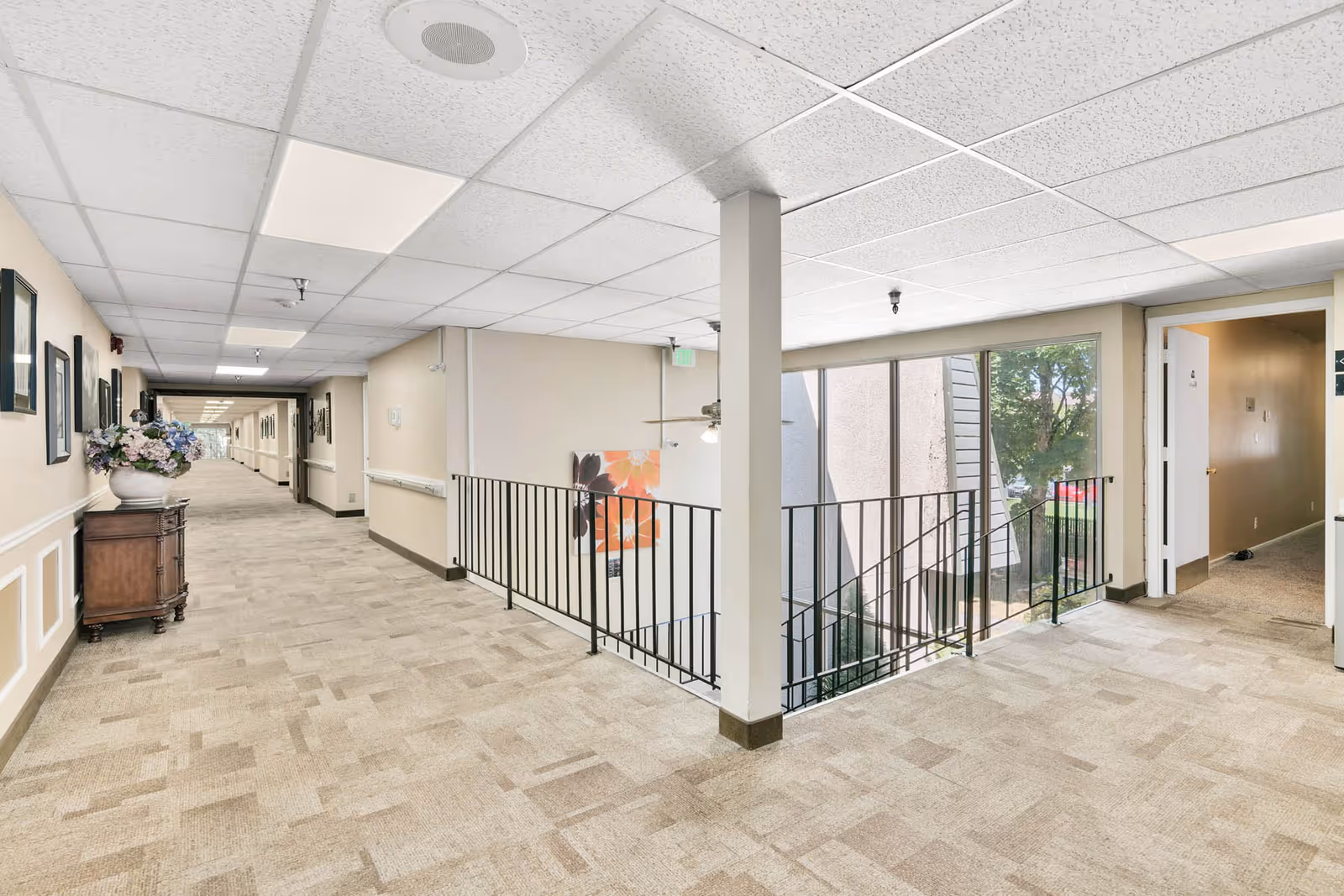 A bright hallway in a senior living facility with beige carpet and walls, decorated with framed pictures and a vase of flowers on a wooden cabinet. The hallway extends into the distance with handrails along the walls. To the right, there is a stairwell with black metal railings and large windows letting in natural light, as well as an open doorway leading to another room.