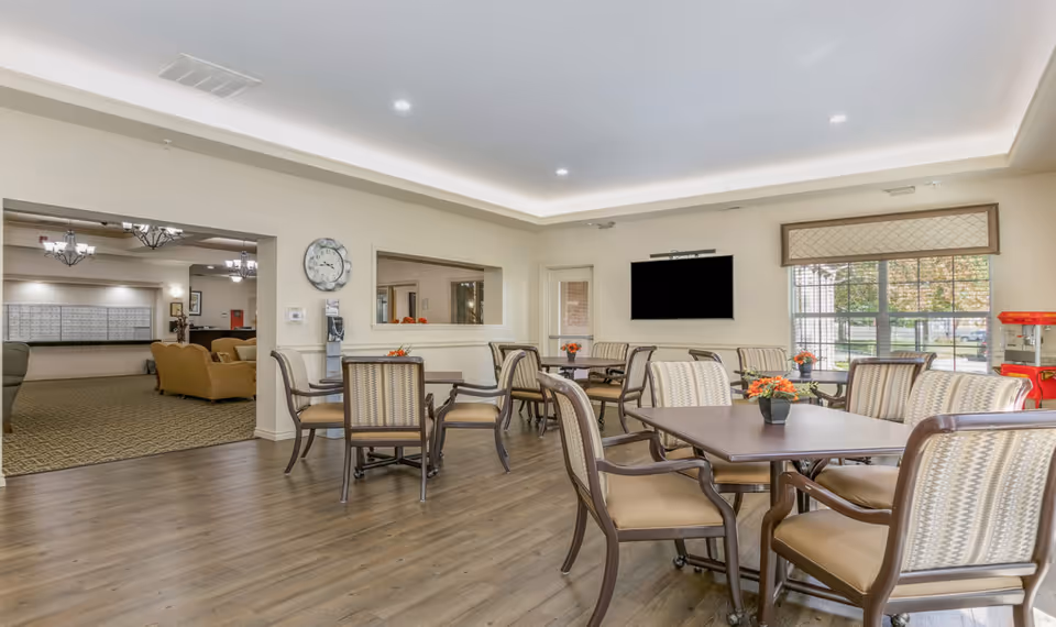 Bright senior community dining/activity room with multiple tables and upholstered chairs, a wall-mounted TV, large windows, and a view into a lounge area.