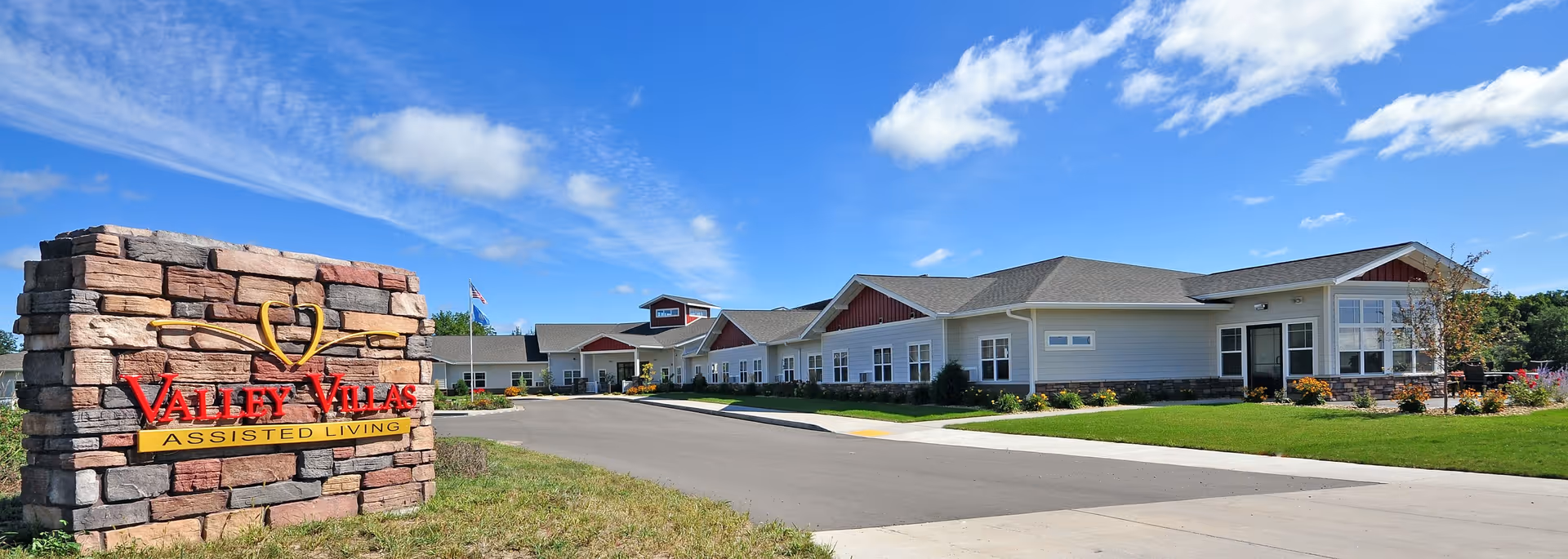 Exterior view of Valley Villas Assisted Living facility on a sunny day with a clear blue sky. The building is a single-story structure with white siding and a gray roof. In the foreground, there is a stone sign with the facility's name and logo. The surrounding area includes a paved driveway, green lawn, and some landscaping.
