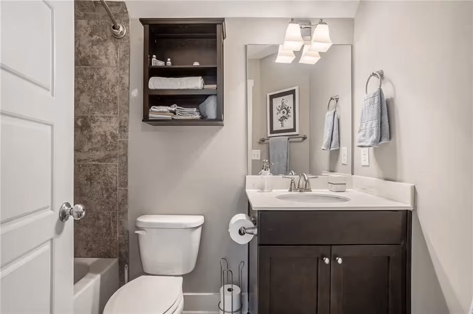 A clean and modern bathroom featuring a white toilet, a dark wood vanity with a white countertop and sink, a large mirror above the sink with a light fixture, a towel ring with a blue hand towel, a wall-mounted cabinet with shelves holding folded towels and toiletries, and a tiled shower area partially visible behind a white door.