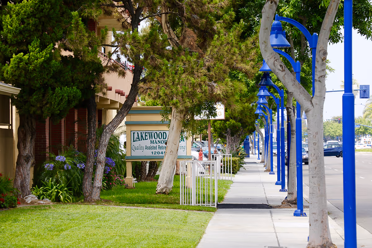 Sidewalk bordered by trees and blue street lamps leading to the Lakewood Manor sign and building entrance.
