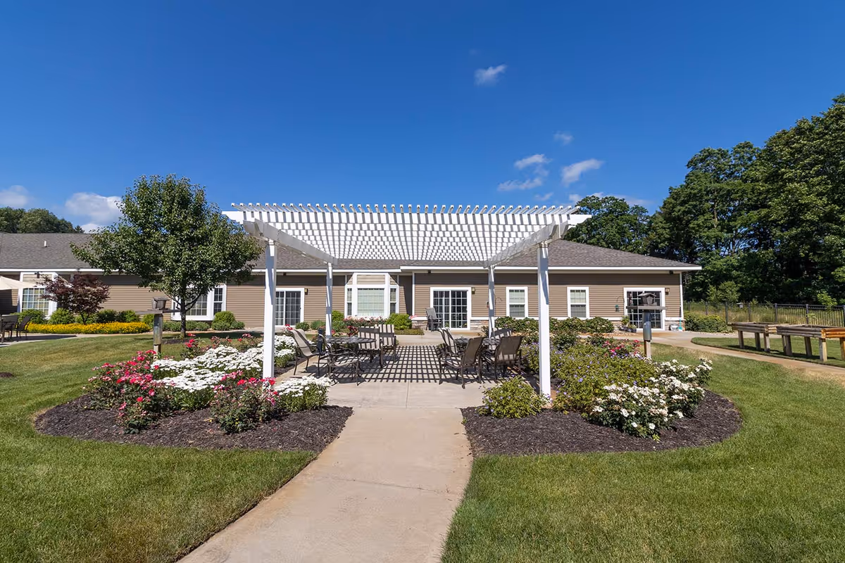 Outdoor courtyard with a white pergola, patio seating and flower beds in front of a single-story assisted living building under a blue sky.