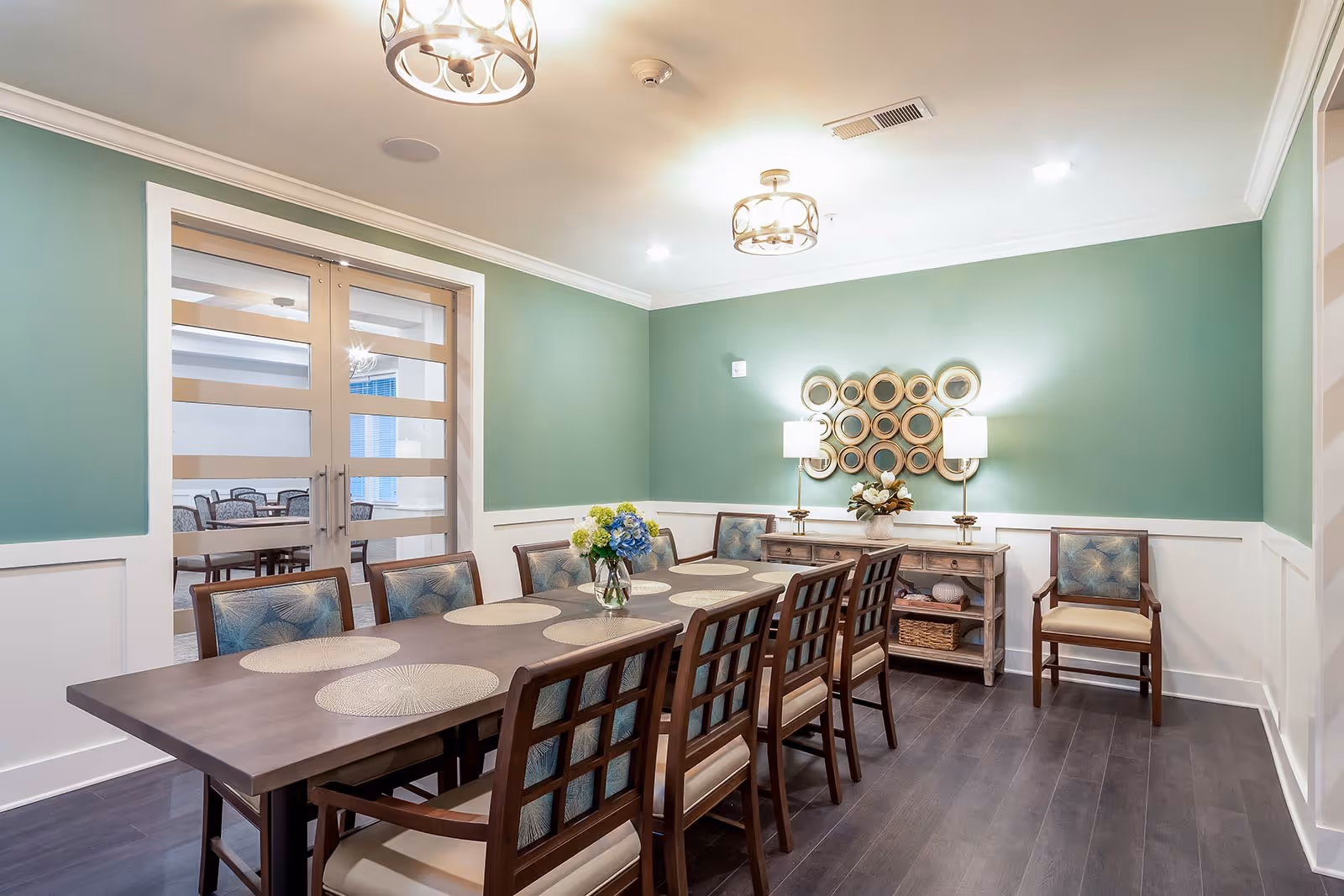 A dining room with a long wooden table surrounded by eight chairs with patterned cushions. The walls are painted green with white wainscoting, and there are two decorative lamps on a sideboard against the wall. A circular mirror wall art piece hangs above the sideboard. Double glass doors lead to another room with more tables and chairs.