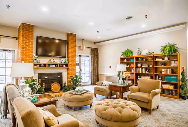 A cozy living room area featuring beige armchairs and tufted ottomans arranged around a brick fireplace with a mounted flat-screen TV above it. The room includes wooden bookshelves filled with books and decorative plants, a side table with a lamp and flowers, and large windows with curtains allowing natural light to enter.