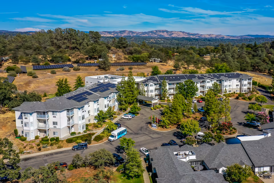 Aerial view of a senior living campus featuring white multi-story buildings with solar panels, parking areas, trees, and distant hills.
