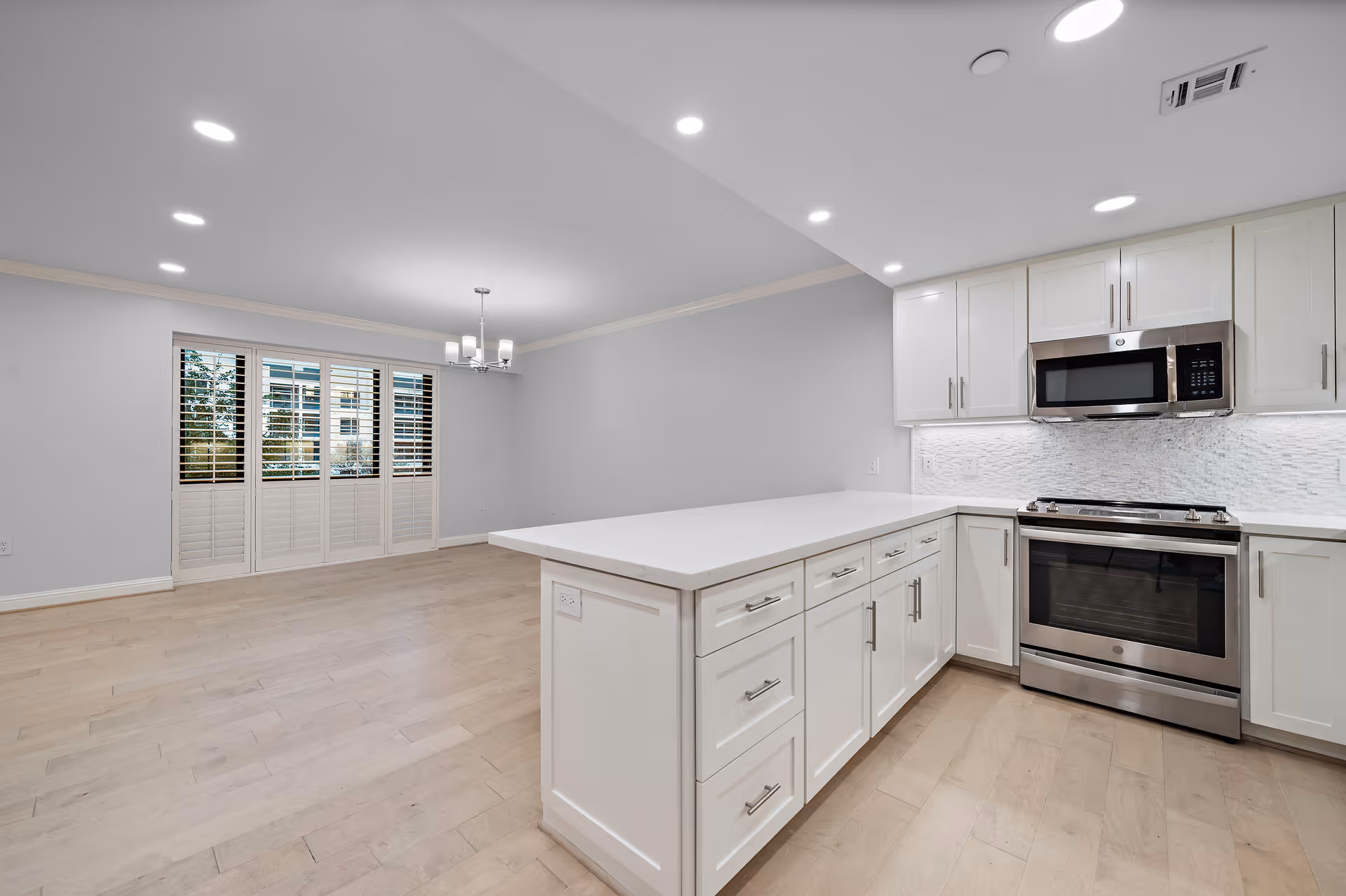 Bright open-concept kitchen with white cabinets, a large island, stainless steel stove and microwave, and an adjoining dining area with shuttered windows.