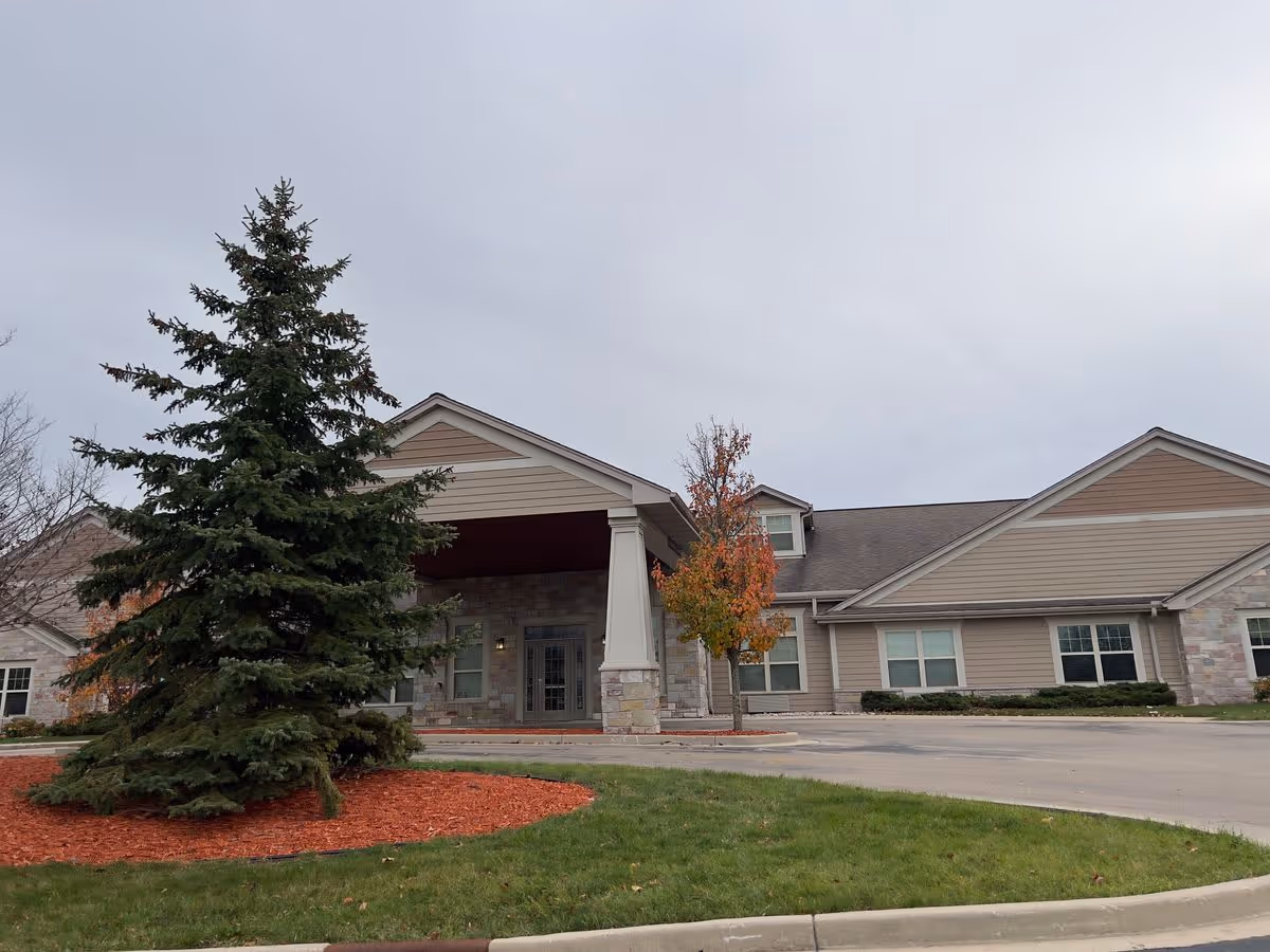Front entrance of a single-story senior living building with a covered portico, an evergreen tree, and landscaped driveway.