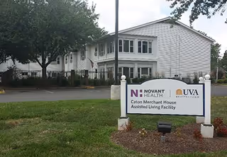 Exterior view of a white two-story assisted living facility building with multiple windows and a parking area in front. A sign in the foreground reads 'Novant Health UVA Health System Caton Merchant House Assisted Living Facility'. Trees and landscaping surround the building.