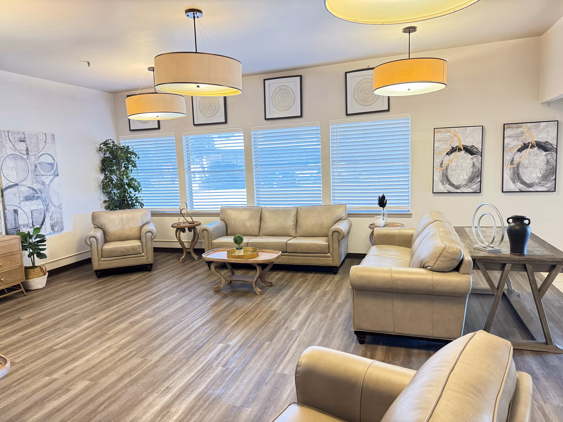 A spacious and well-lit living room with beige leather sofas and armchairs arranged around wooden coffee tables. The room features large windows with blinds, modern pendant lights hanging from the ceiling, framed abstract artwork on the walls, and decorative plants in the corners.