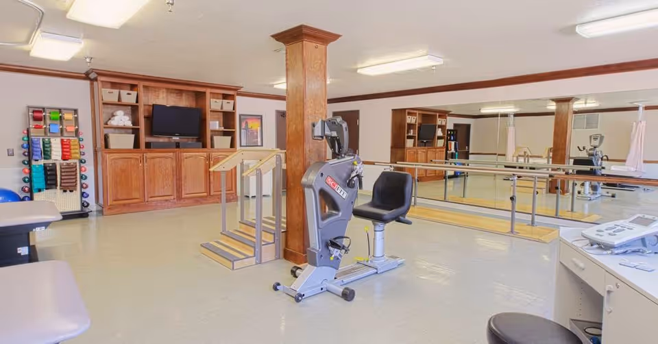 A spacious physical therapy or exercise room with light-colored flooring and walls. The room features a recumbent exercise bike, parallel bars for walking support, a rack with various colored dumbbells and exercise bands, and wooden cabinetry with a television and storage baskets. Large mirrors cover one wall, reflecting the equipment and room interior. The space is well-lit with ceiling lights and has a clean, organized appearance.