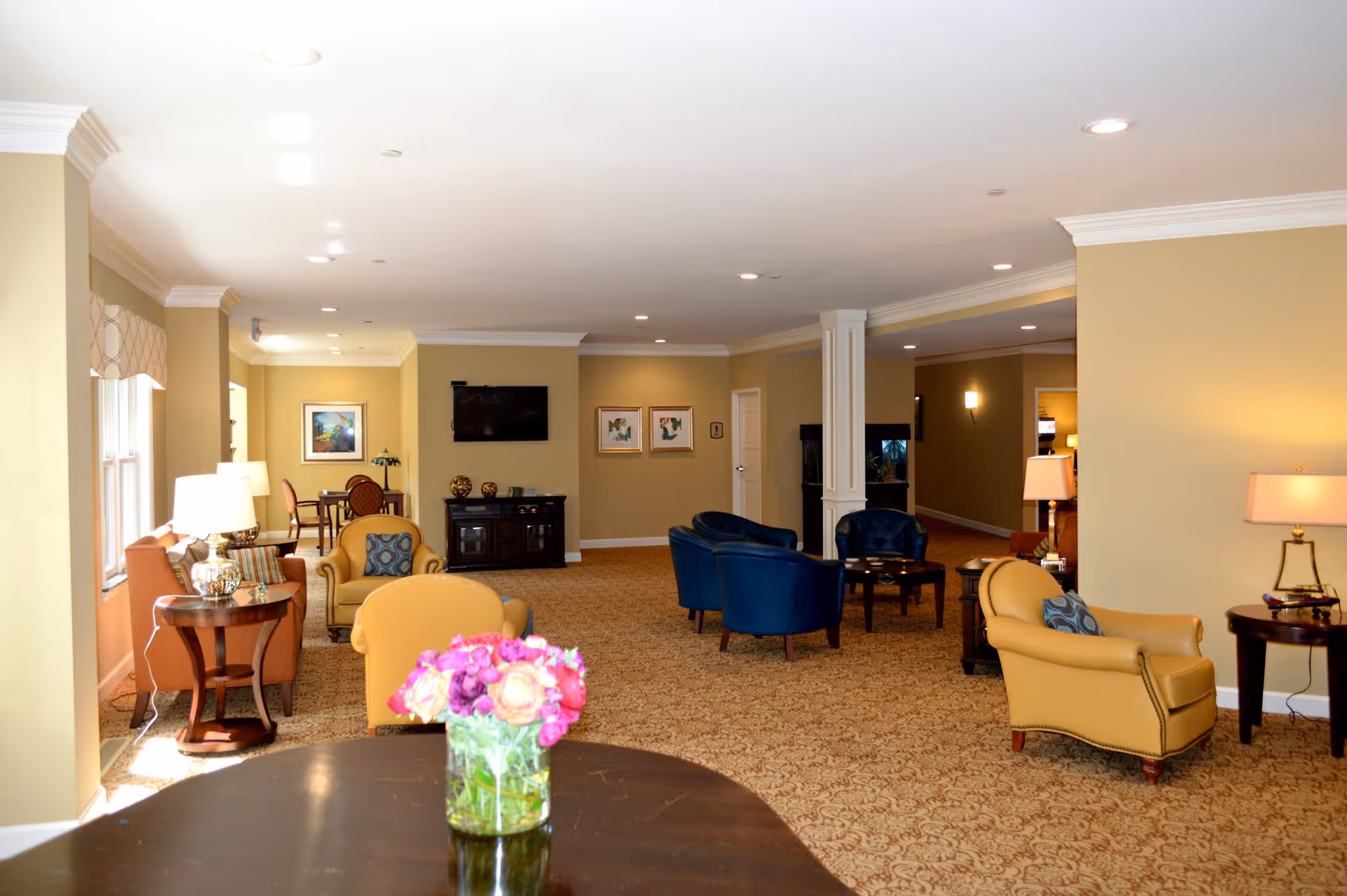 A spacious and well-lit living room area in a senior living facility with beige walls and carpeted floor. The room features several comfortable armchairs in yellow and blue, side tables with lamps, a flat-screen TV mounted on the wall, framed artwork, and a small table with a vase of colorful flowers in the foreground.