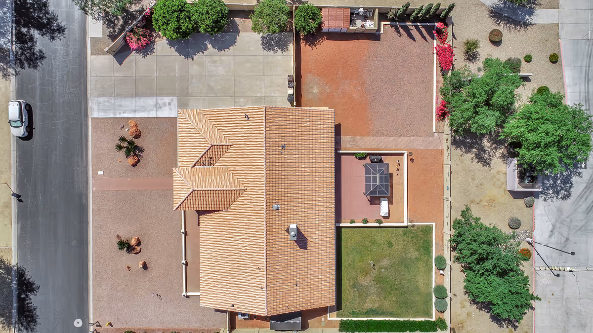 Aerial view of a residential property with a tiled roof, surrounding yard areas including a green lawn, a patio with a gazebo, gravel sections, and landscaped plants. The property is bordered by streets and sidewalks.