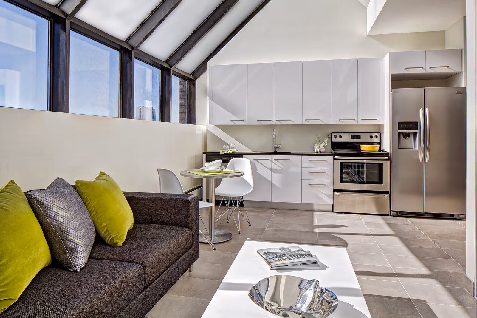 Modern open-concept living area with a dark gray sofa adorned with green and gray pillows, a small round dining table with two white chairs, and a kitchen featuring white cabinets, a stainless steel refrigerator, and an oven. Large windows with black frames allow natural light to fill the space.