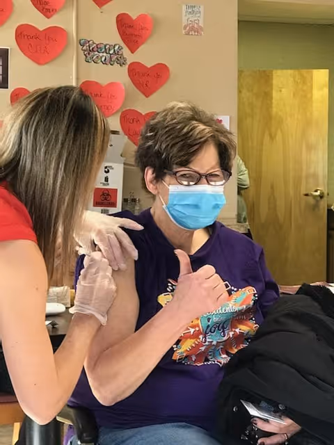 A woman wearing glasses and a blue face mask is sitting and giving a thumbs-up while receiving a vaccine injection in her upper arm from a healthcare worker wearing gloves. The background shows a wall decorated with red heart-shaped notes and a 'Thank You' sign.