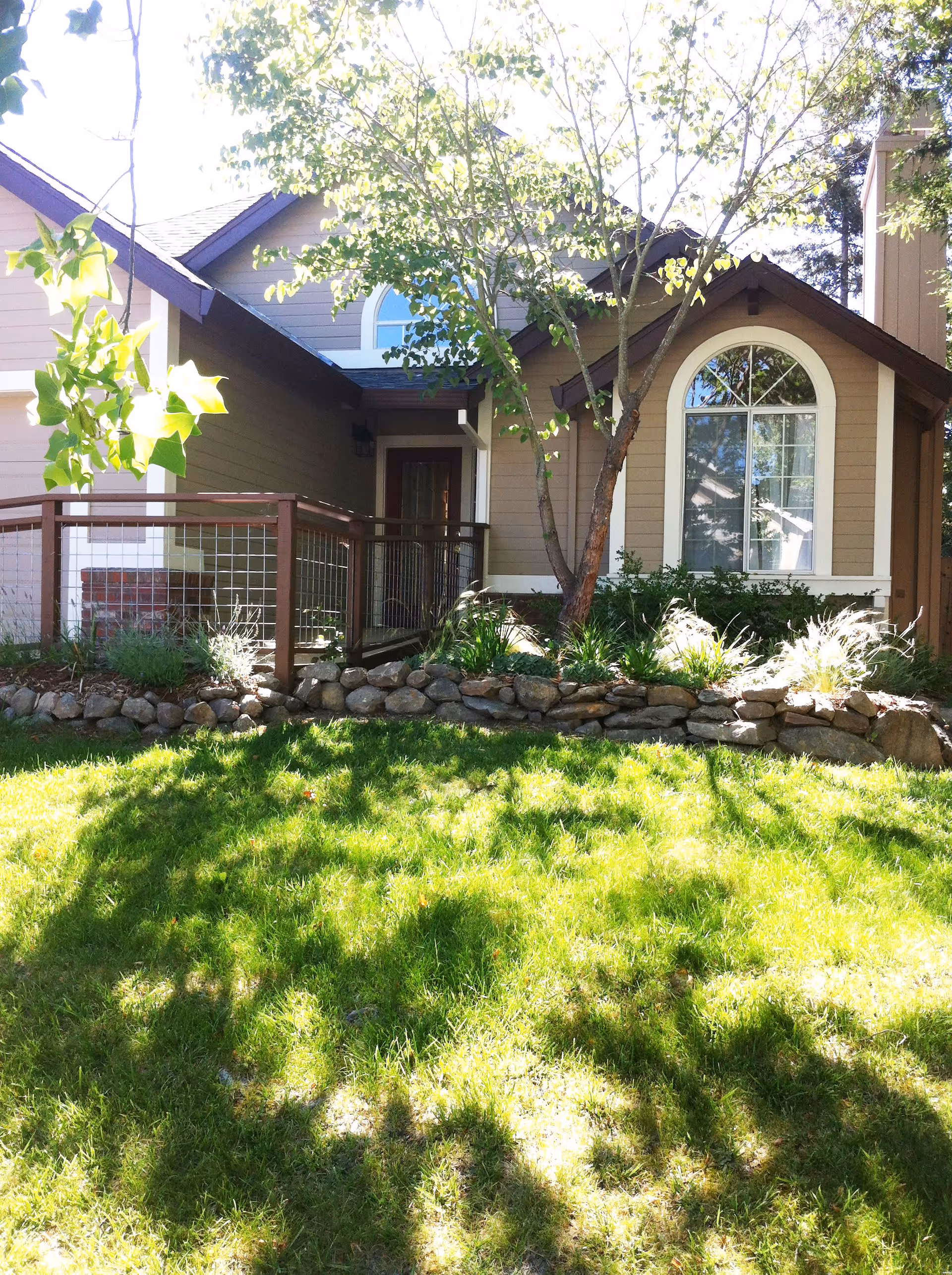 Front exterior view of a single-story house with beige siding, a brown roof, and a large arched window. There is a small porch with a wooden railing and a tree in front of the house casting shadows on the green lawn.