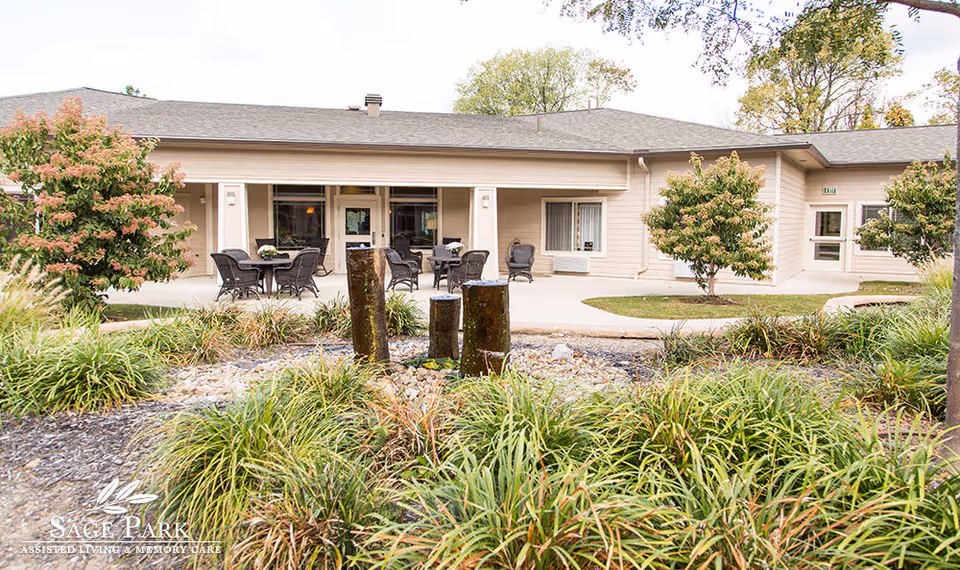 Front exterior of a single-story assisted living building with patio seating, landscaping, and a small water feature.