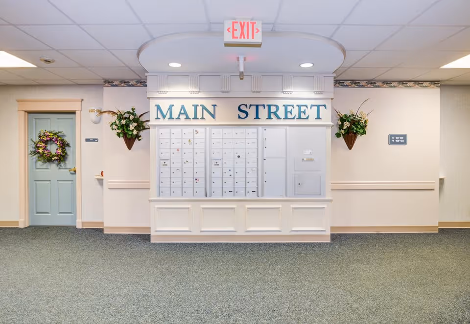 Interior hallway area with a wall of mailboxes labeled 'MAIN STREET' above them. There is a light blue door decorated with a floral wreath on the left side and two wall-mounted flower arrangements on either side of the mailboxes. An exit sign is mounted on the ceiling above the mailboxes.