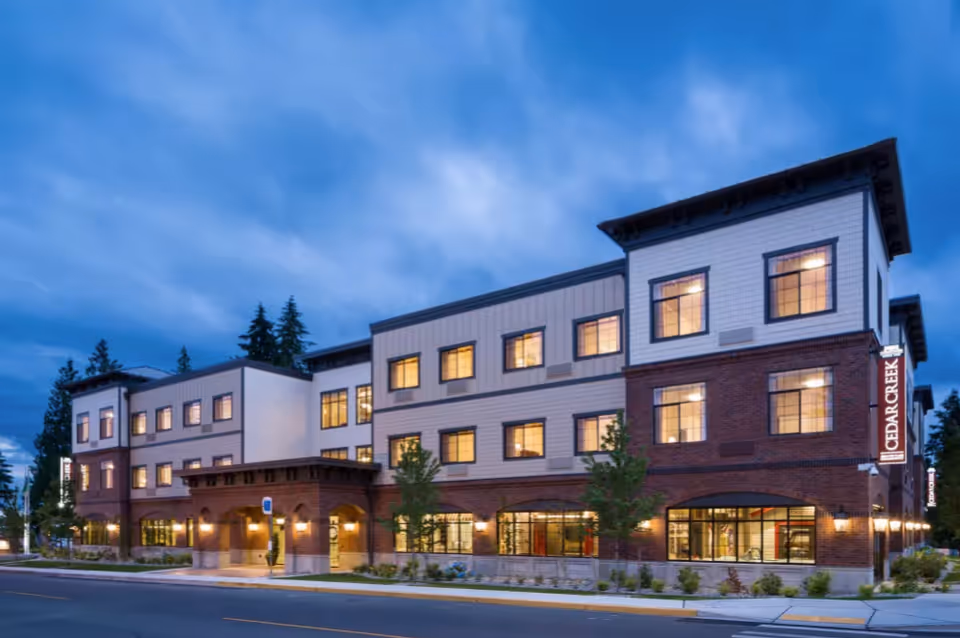 Exterior view of a three-story building at dusk with lights on inside. The building has a combination of brick and white siding with multiple windows and a sign that reads 'Cedar Creek'. Trees and landscaping are visible around the building along a sidewalk and street.