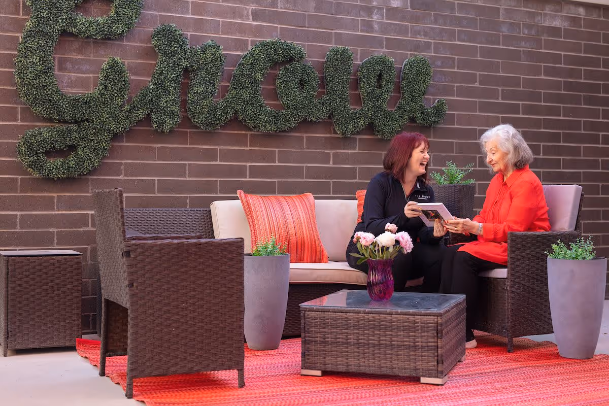 Two women sitting and smiling on a wicker outdoor sofa with red cushions, holding a booklet. They are on a red rug with a wicker coffee table holding a purple vase with flowers. Behind them is a brick wall with green shrubbery shaped into the word 'groove'.
