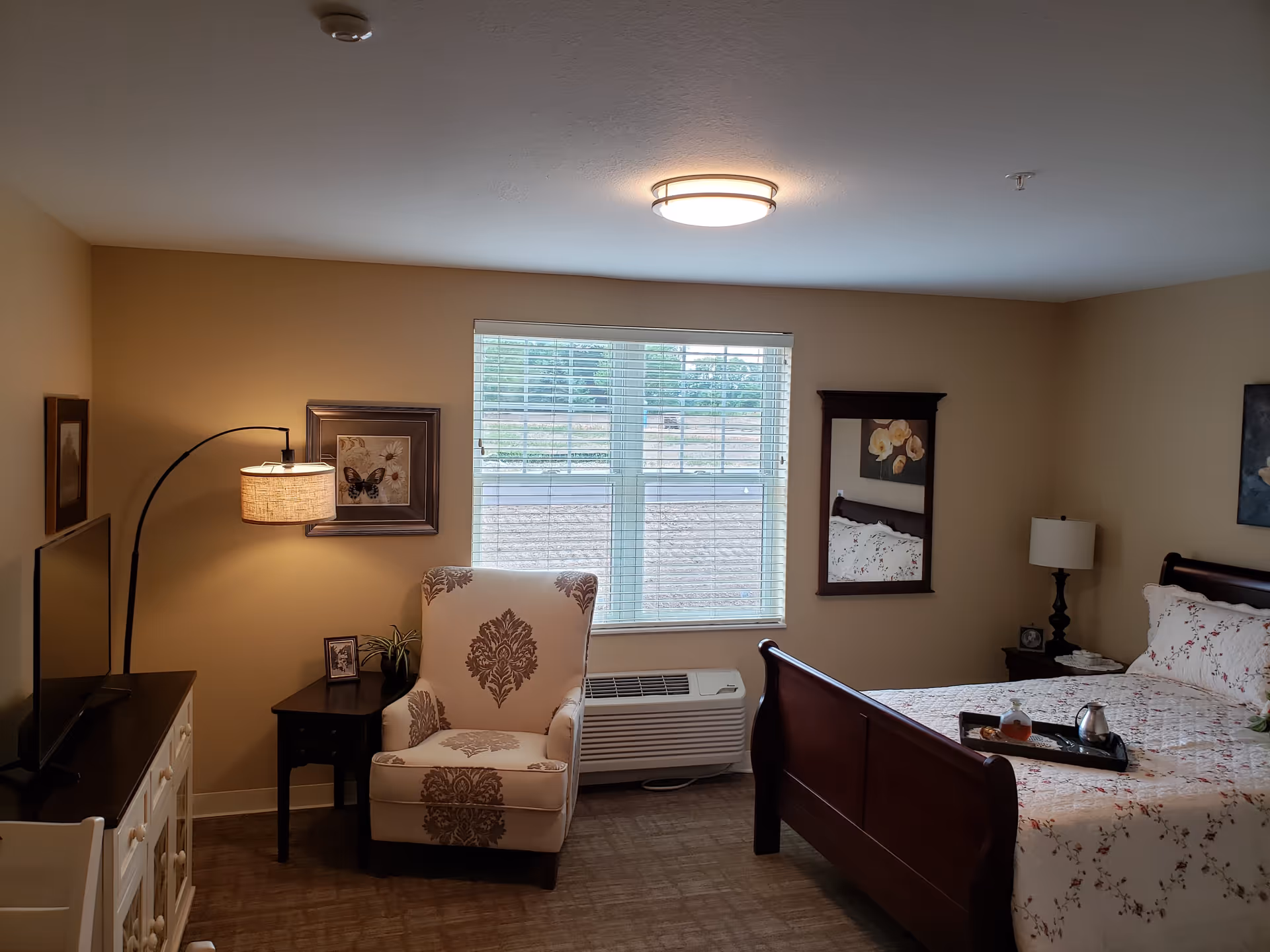 A cozy bedroom with beige walls and carpeted floor. The room features a wooden bed with floral bedding, a tray with a teapot and cup on the bed, a patterned armchair next to a small dark wooden side table with a plant and framed photo, a floor lamp with a beige shade, a flat-screen TV on a white cabinet, a window with blinds, a wall mirror, and framed artwork on the walls.