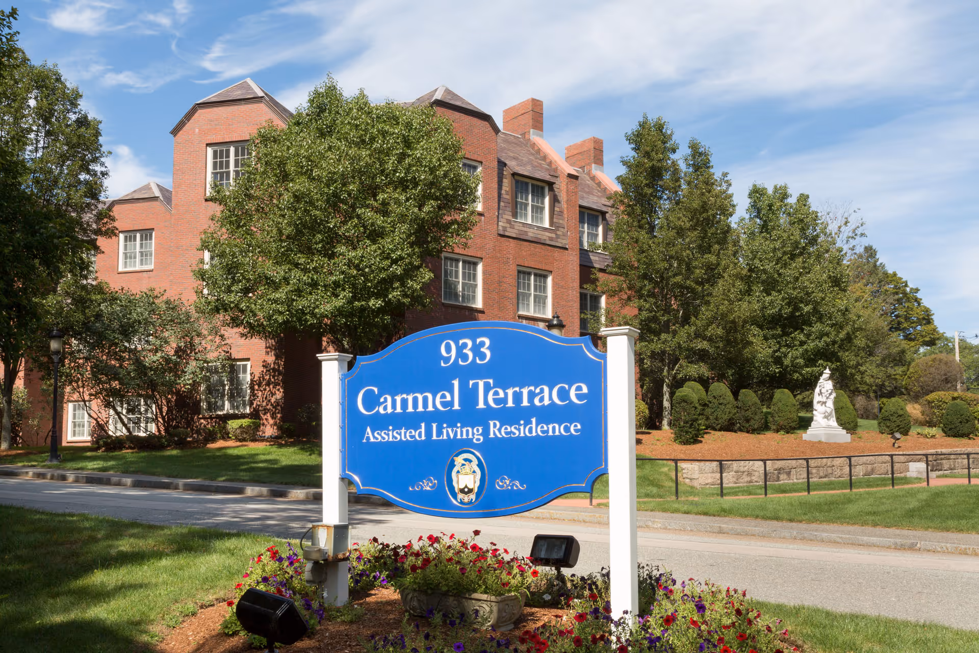 A blue sign with white text reading '933 Carmel Terrace Assisted Living Residence' is displayed in front of a red brick building surrounded by green trees and landscaping under a partly cloudy sky.