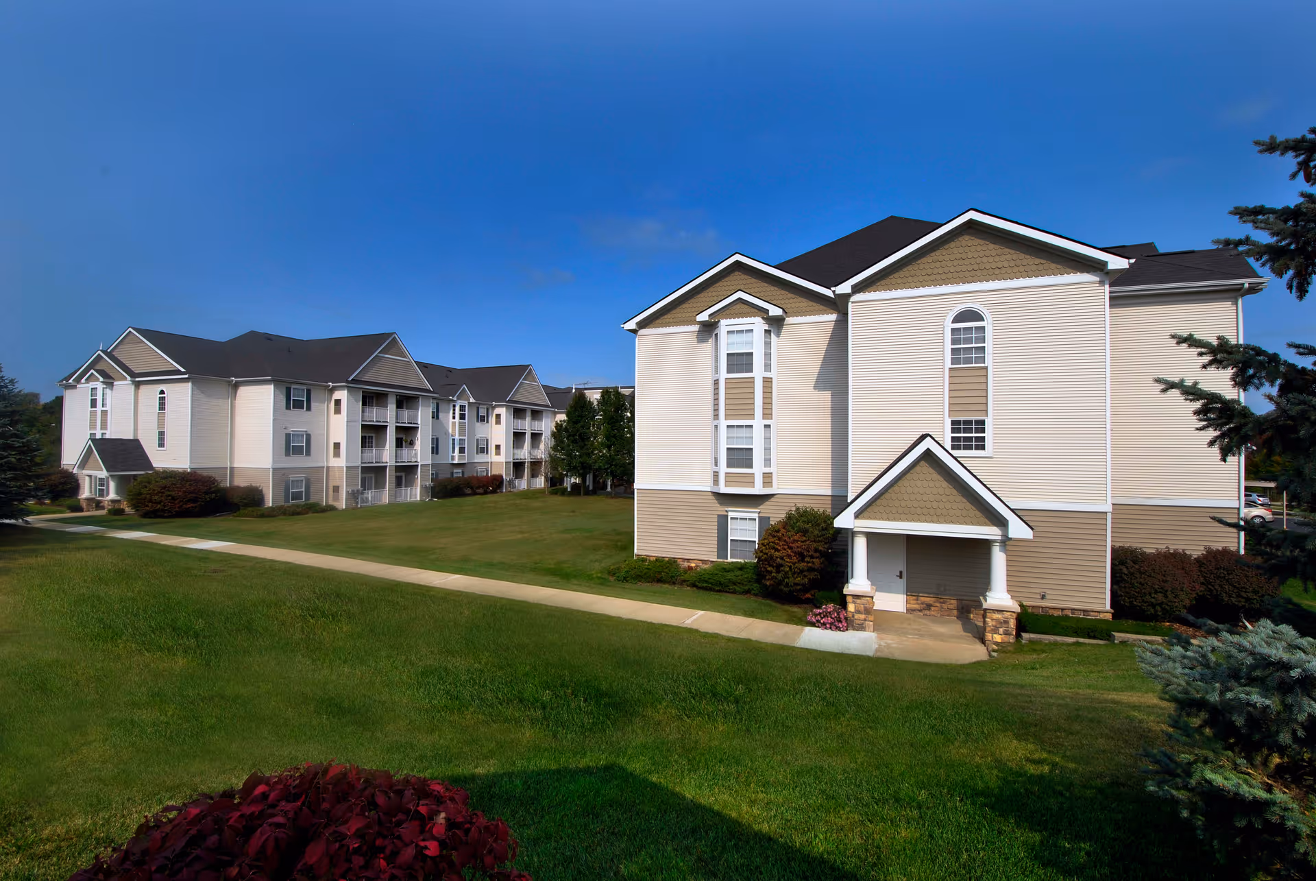 Exterior view of Oakhaven Manor, showing two beige multi-story residential buildings with white trim, surrounded by green lawns and trees under a clear blue sky.