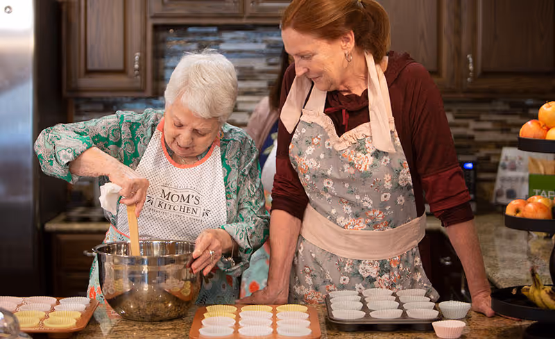 Two elderly women wearing aprons are in a kitchen preparing to bake. One woman is stirring ingredients in a large metal mixing bowl, while the other watches and smiles. There are trays with cupcake liners on the counter in front of them, and kitchen cabinets and appliances are visible in the background.