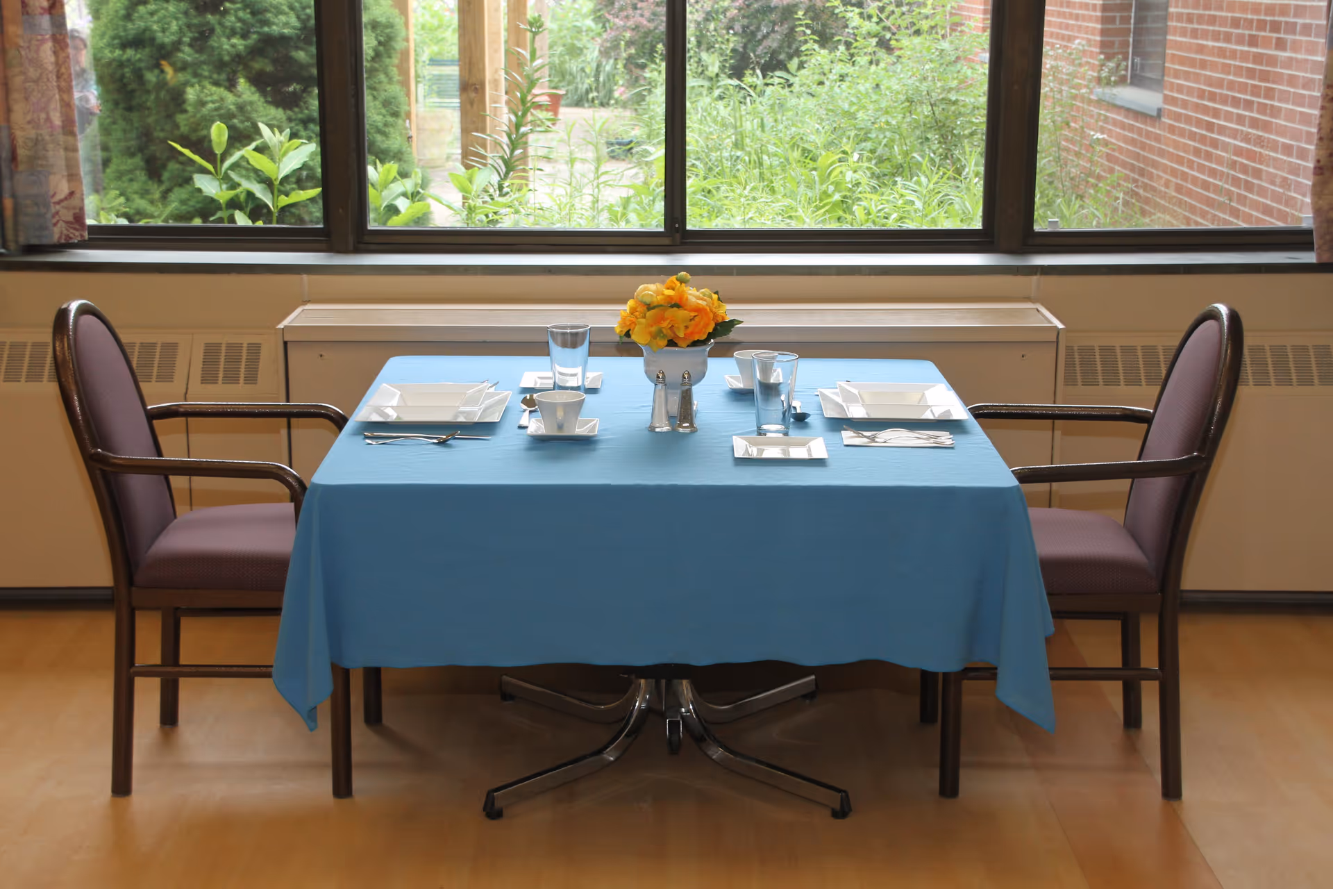 A dining table set for two with a blue tablecloth, white square plates, cups, glasses, silverware, and a small flower arrangement in the center. Two chairs with armrests are positioned on either side of the table. A large window behind the table shows green plants and a brick wall outside.