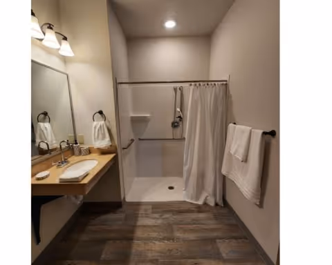 A clean and modern bathroom with a walk-in shower featuring a white curtain, a built-in shower seat, and grab bars. There is a wooden countertop with a white sink, a mirror above it, and two wall-mounted light fixtures. White towels hang on black towel rings and a towel bar. The floor has wood-like tiles.