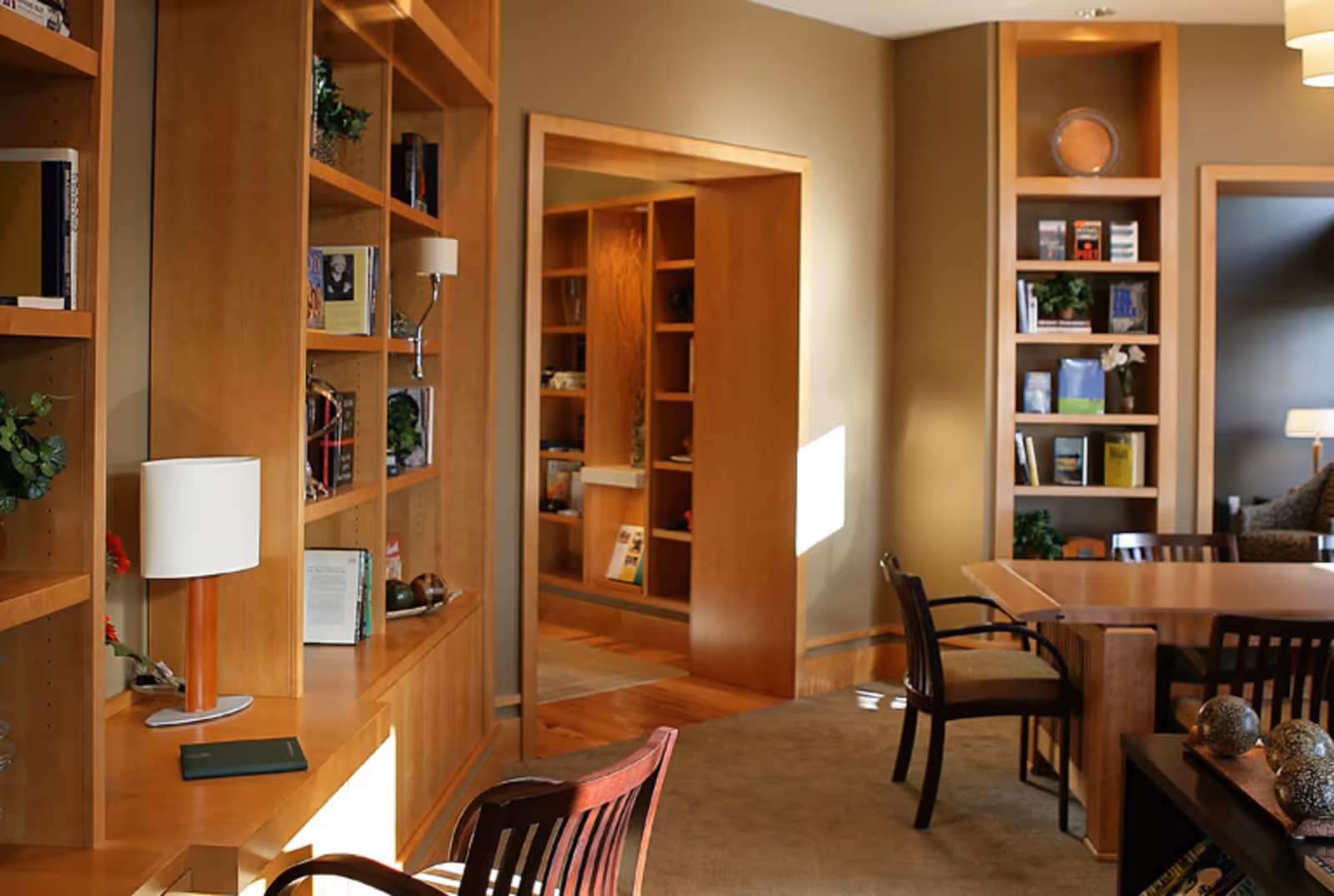 A cozy interior room with wooden bookshelves filled with books and decorative items. There is a wooden table with chairs around it, a lamp on a side table, and a doorway leading to another room with more bookshelves. The room is warmly lit with natural light coming through a window.