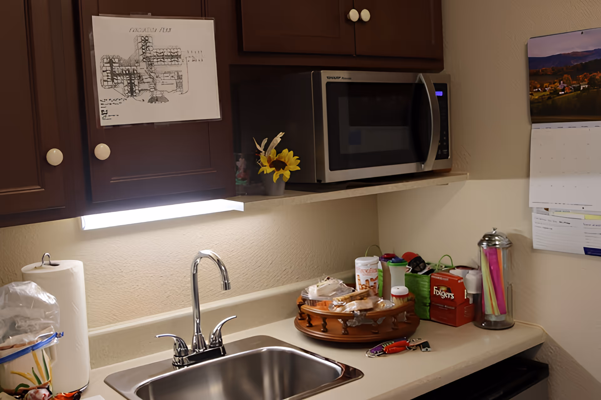 A kitchen countertop with a stainless steel sink and faucet, a paper towel roll, a microwave oven on the counter, a small vase with yellow flowers, a rotating tray with snacks and containers, a container with colorful straws, and a wall calendar.