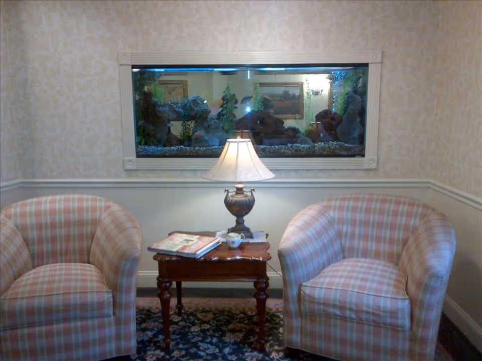 A cozy sitting area with two plaid upholstered armchairs facing a small wooden table with a decorative lamp and some books. Behind the table is a large built-in aquarium with rocks and plants, set into a wall with light patterned wallpaper and white wainscoting.